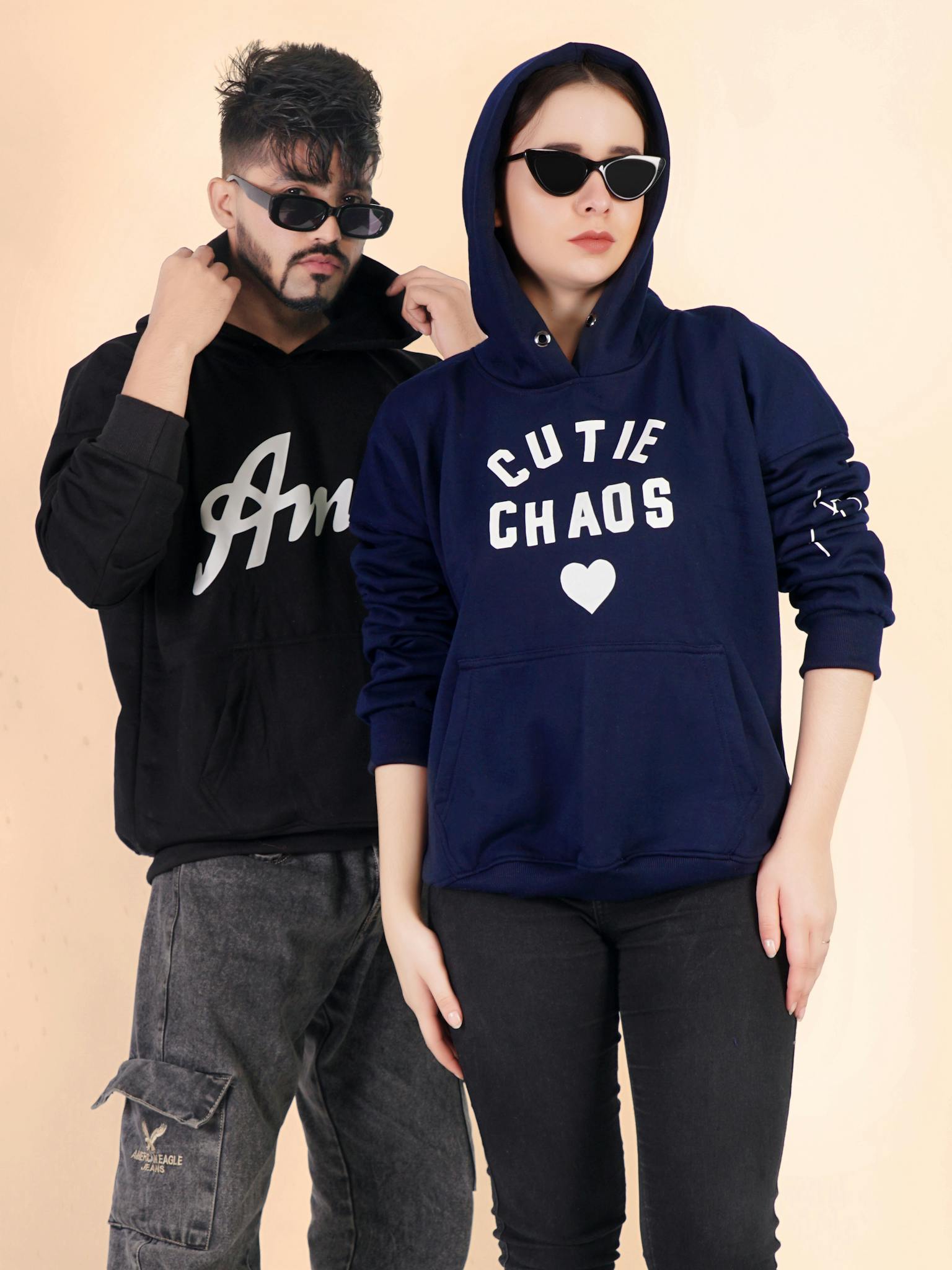Two models in stylish hoodies pose with sunglasses in a studio setting.
